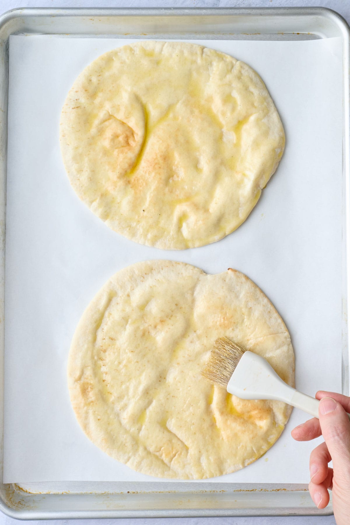 Brushing oil on pita bread on a parchment lined sheet pan.
