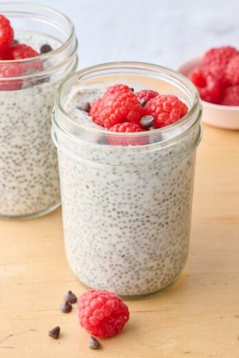 2 Yogurt chia pudding in jar, focus on one, topped with fresh diced strawberries with small spoon nearby, extra fresh fruit nearby.