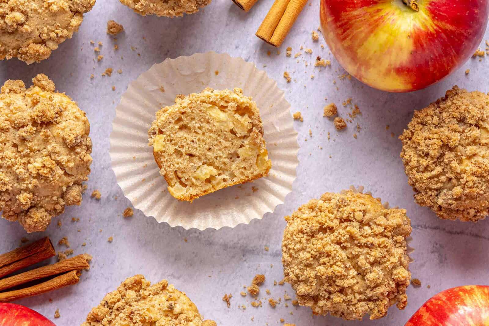 Apple muffins on surface, one with parchment paper pulled off and half of the muffin resting cut side up on it to show inside texture.