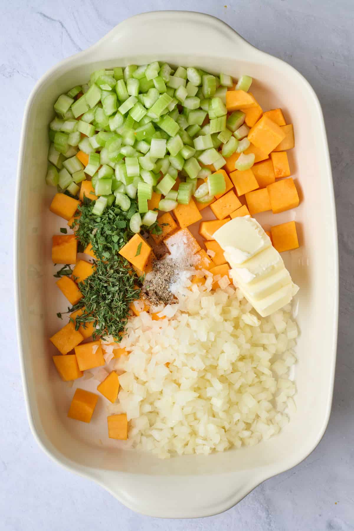 Squash, onion, celery, butter, and seasonings in a baking dish before tossing together.