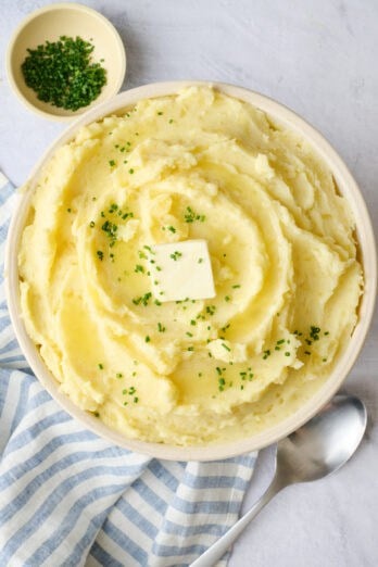 Creamy mashed potatoes in a serving bowl with a pat a mostly melted butter on top, garnished with chopped chives, serving spoon nearby.