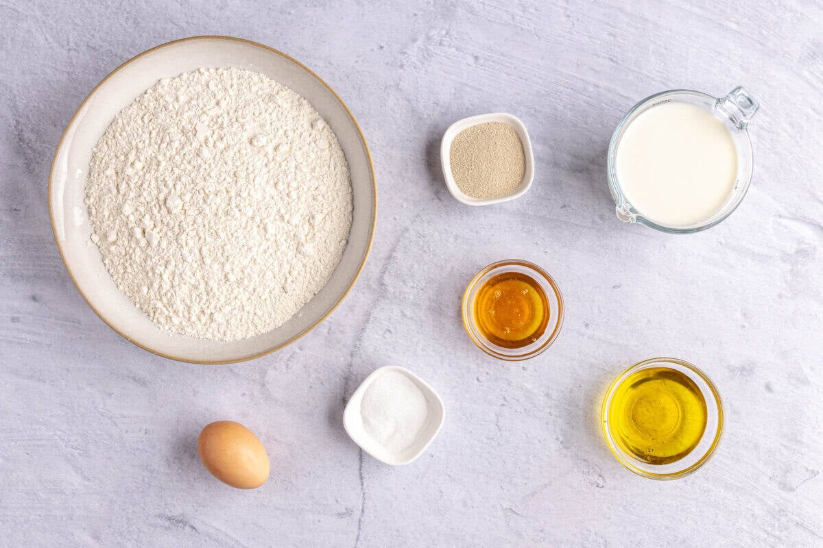 Ingredients for dinner roll recipe in individual dishes: flour, yeast, egg, salt, honey, olive oil, and milk.