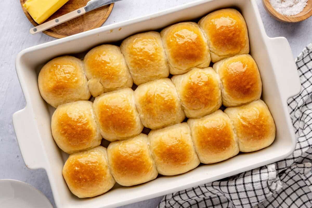 Easy homemade dinner rolls in a baking dish brush with olive oil with butter and flaky salt nearby.