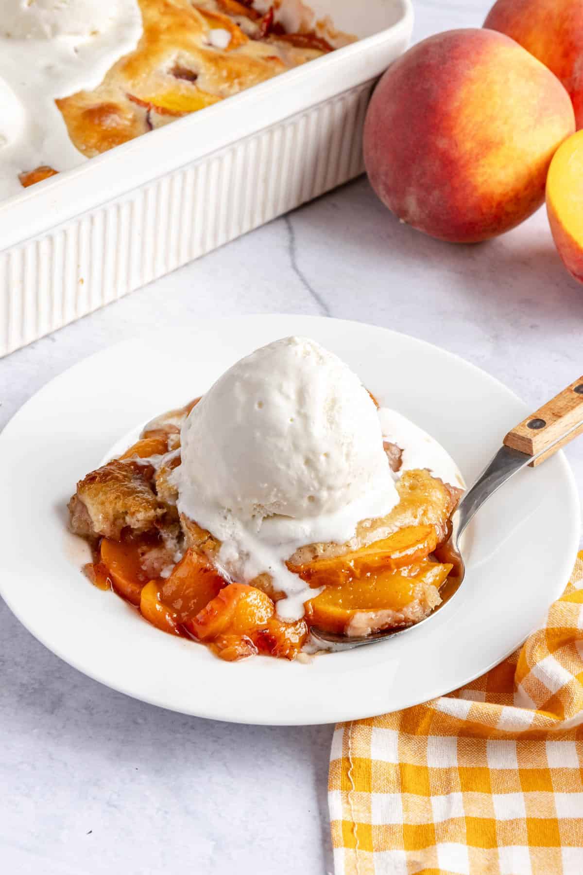 Serving of peach cobbler in a bowl with a few visible slices of peaches. Topped with a scoop of ice cream, baking dish nearby.