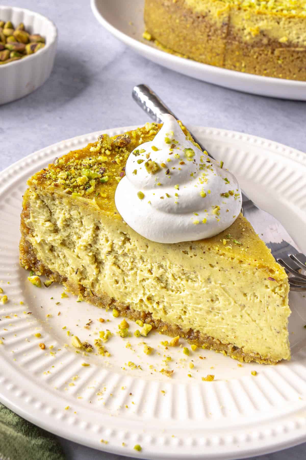 Slice of pistachio cheesecake on a plate topped with a dollop of whipped cream, fork on plate, whole cheesecake nearby.