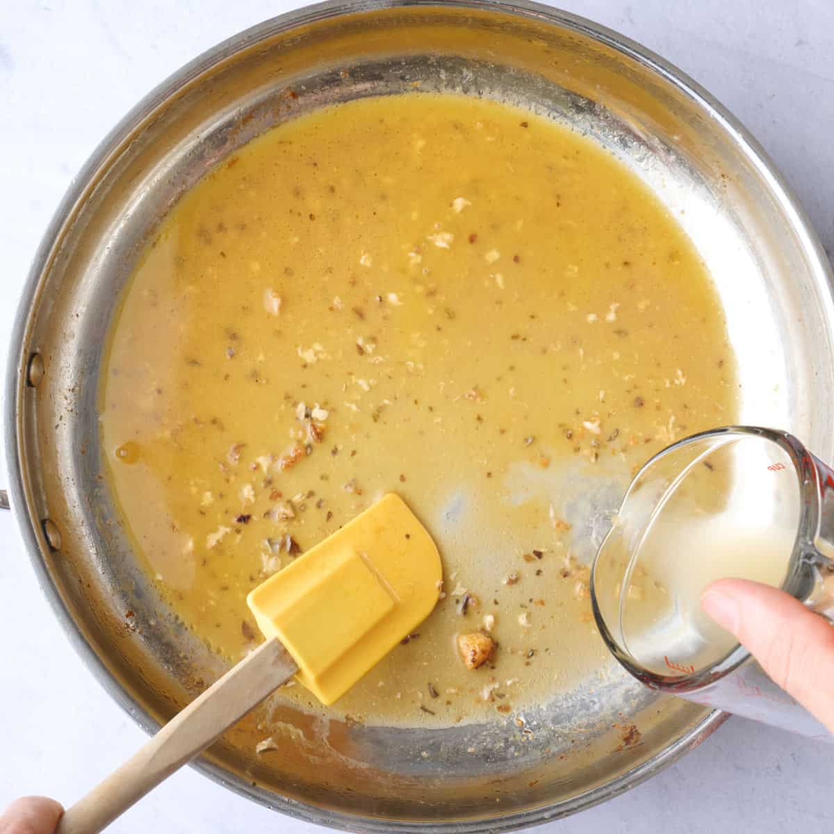 Broth being poured into a seasoned skillet with a rubber spatula lifting up the chicken juice and bits from the bottom of the pan.
