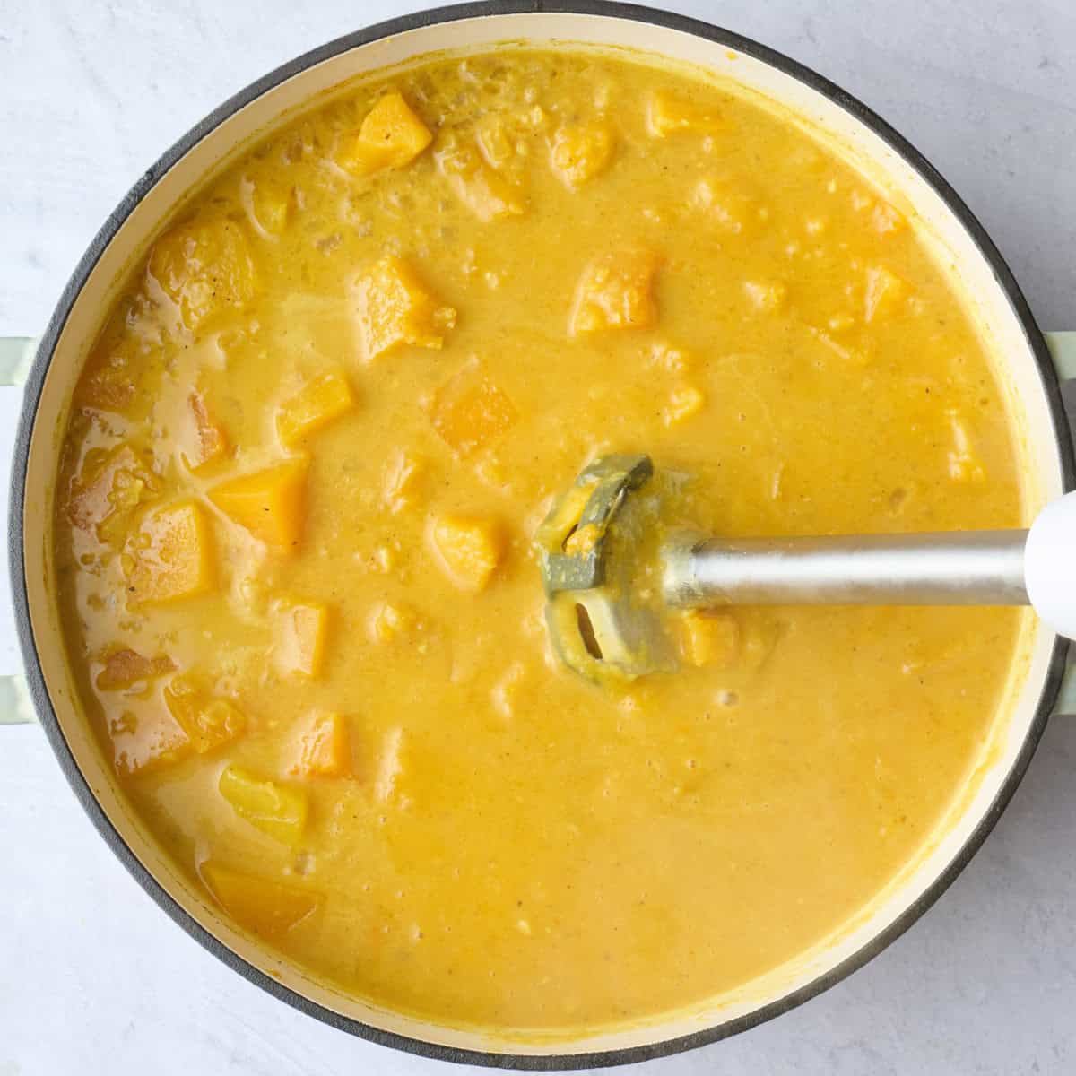 An immersion blender dipped into pot blending up the curried butternut squash soup into a smooth consistency.