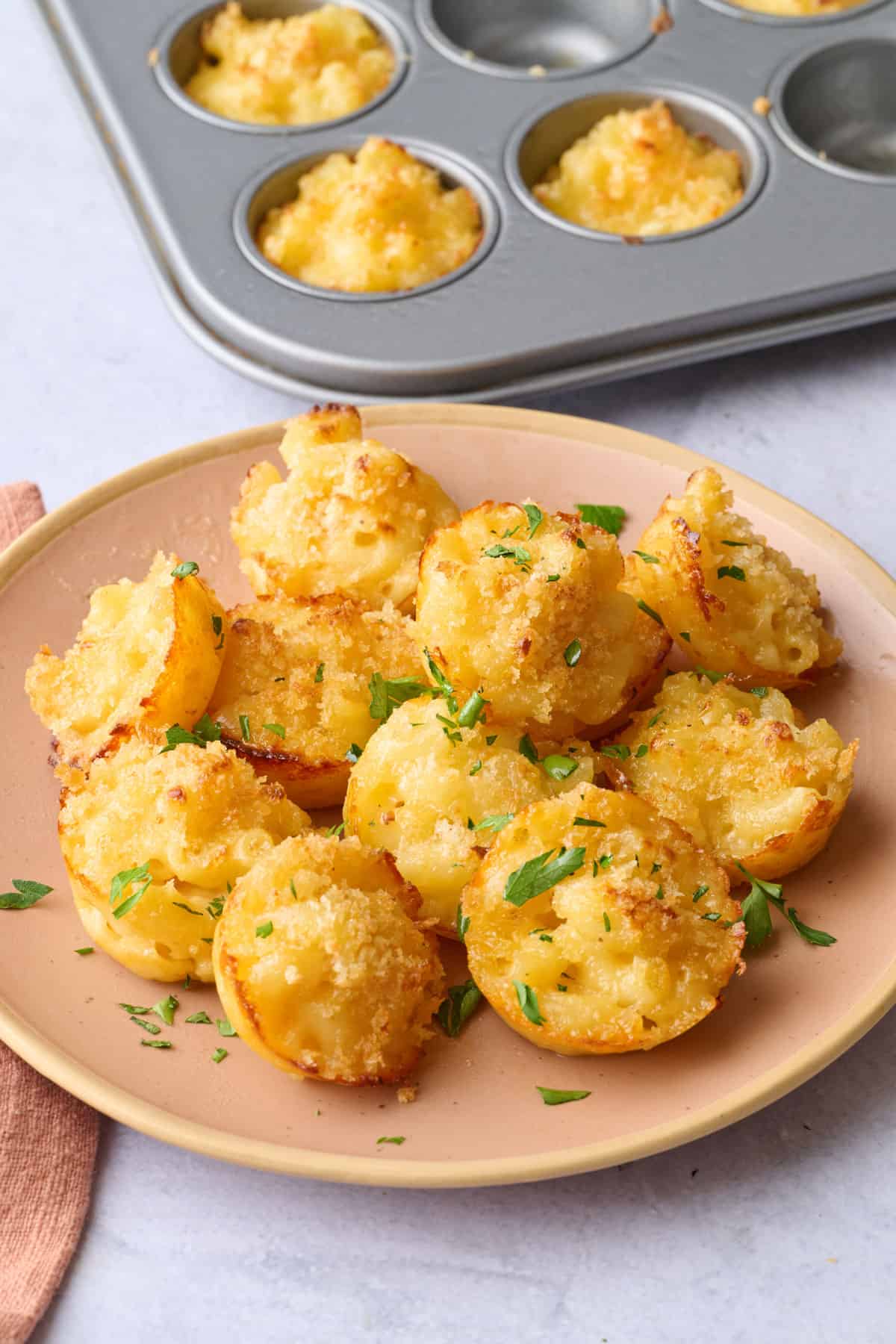 Baked mac and cheese bites on a plate with mini muffin pan of more nearby.