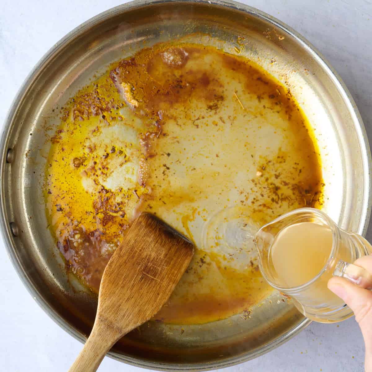 Stock being poured into a skillet with a wooden spoon, scraping up browned bits from cooking chicken.