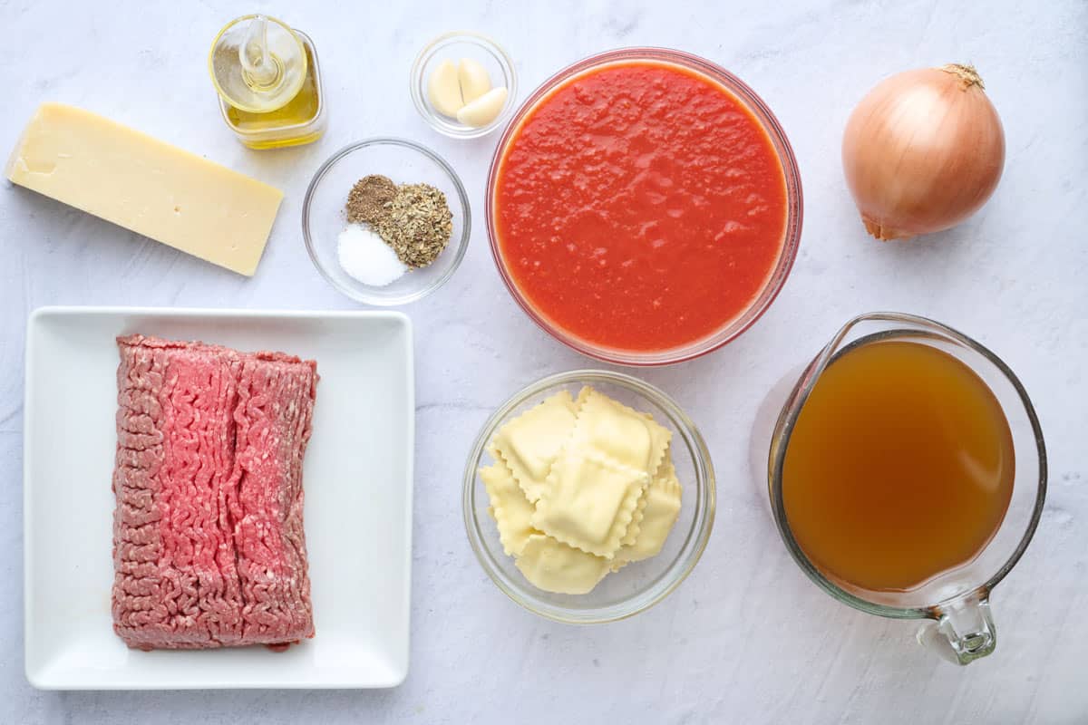 Ingredients for recipe: lean ground beef: parmesan cheese block, oil, seasonings, garlic cloves, crushed tomatoes, ravioli, onion, and broth.