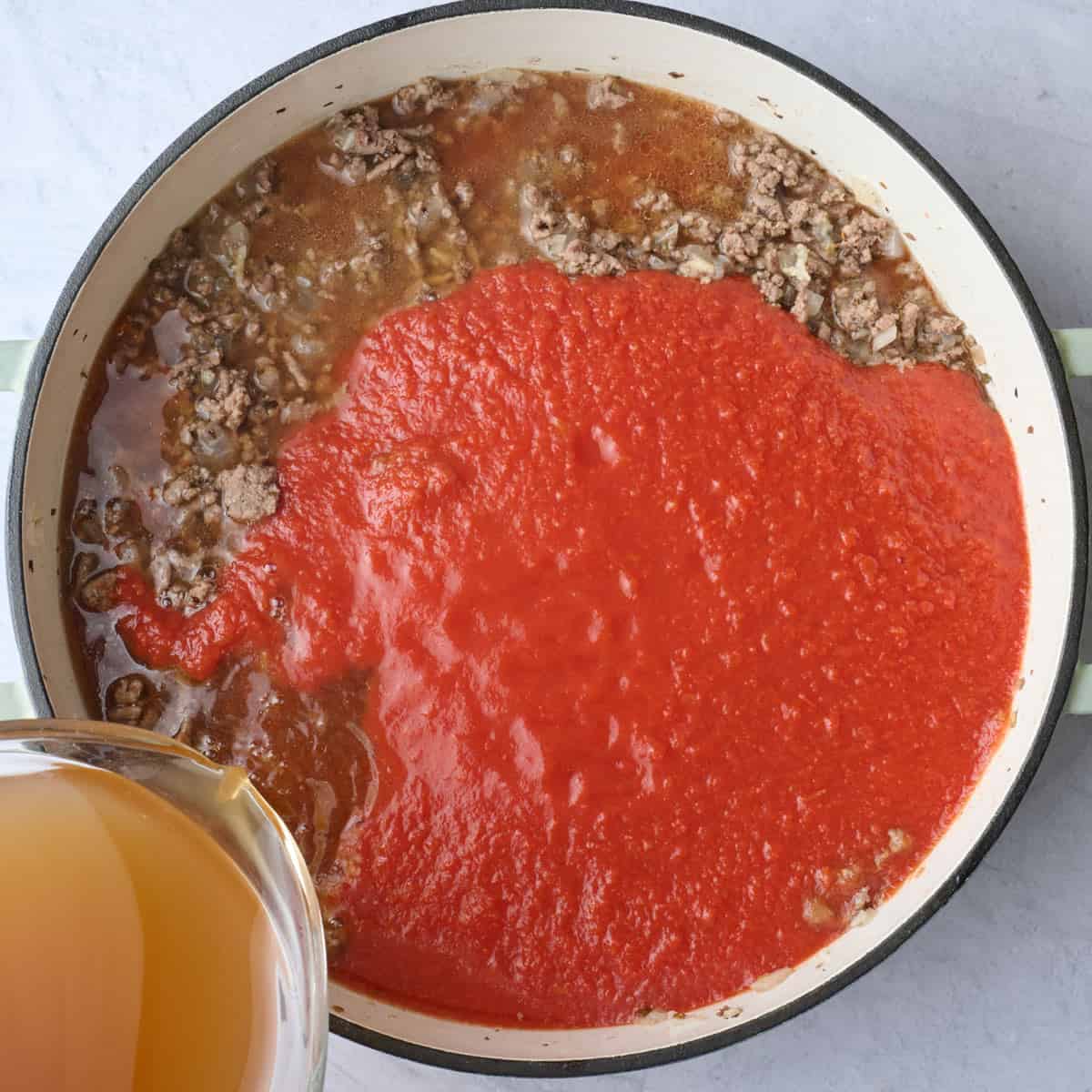 Vegetable broth being poured into a pot of cooked ground beef and onions with crushed tomatoes added before mixing together for soup recipe.