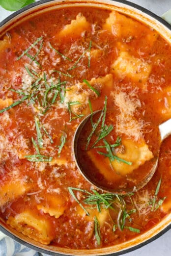 Ravioli soup recipe in a pot with a ladle dipped inside, garnished with parmesan cheese and sliced basil.
