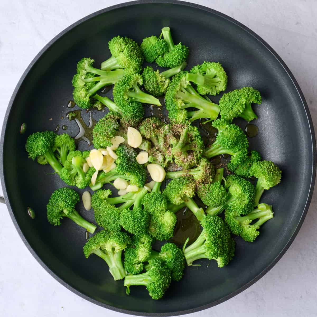 Steamed broccoli florets in a pan with oil, sliced garlic, salt and pepper added on top before cooking.