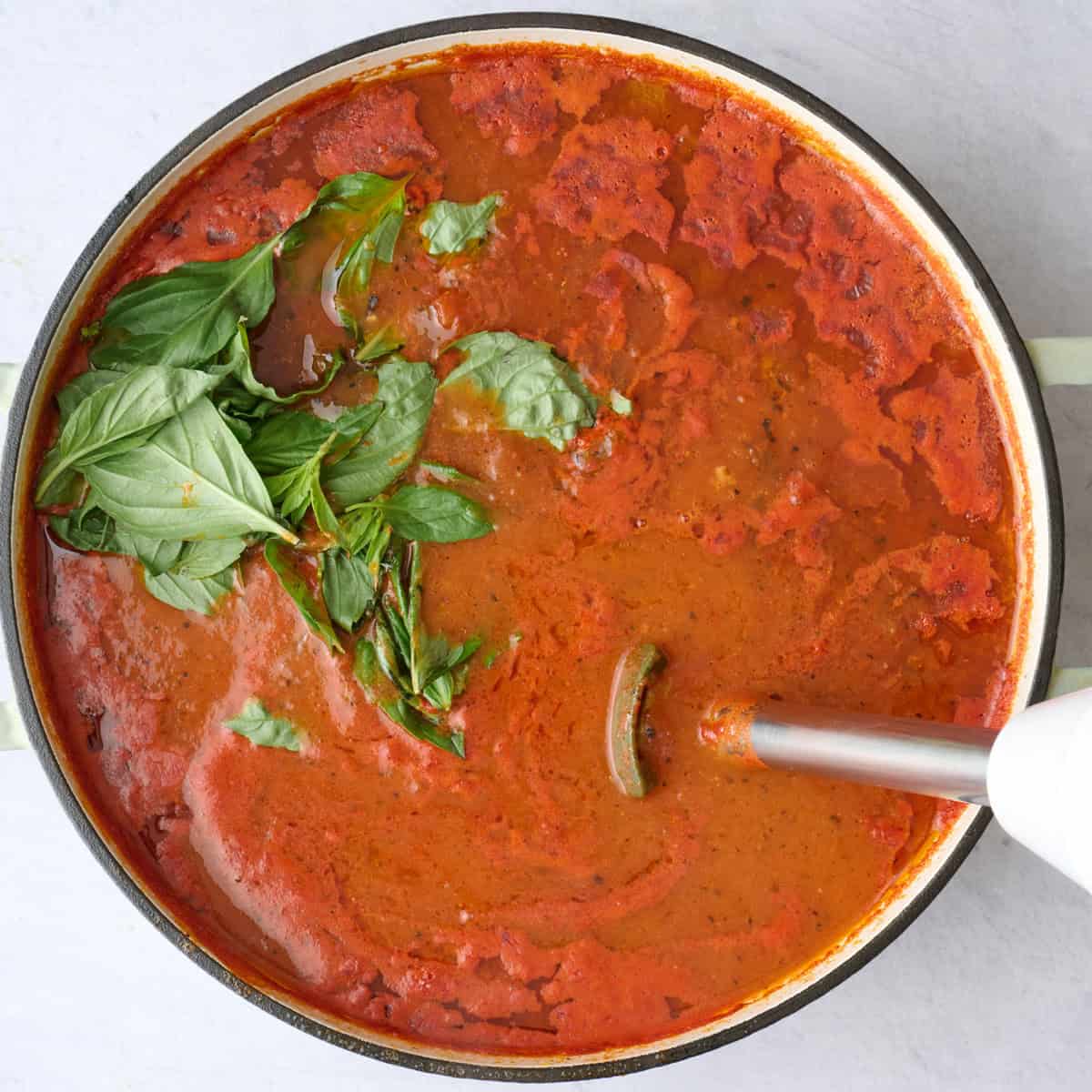 Tomato soup after cooking with fresh basil leaves added to pot and an immersion blender dipped inside before blending.