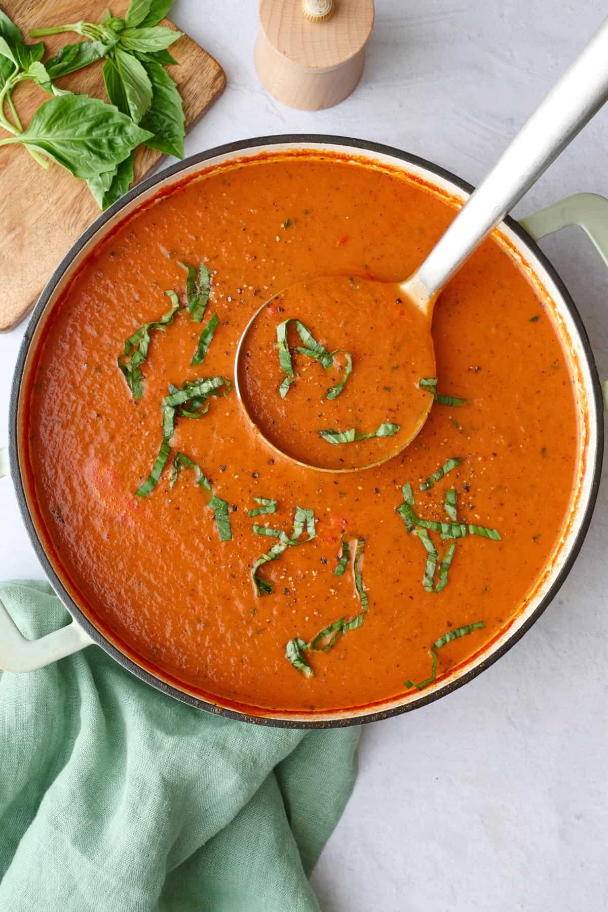 Thick homemade tomato soup in a pot garnished with fresh basil and a ladle lifting some up.