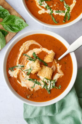 Two bowls of homemade tomato soup garnished with a few croutons and cream swirl, and basil leaves with a spoon dipped inside.