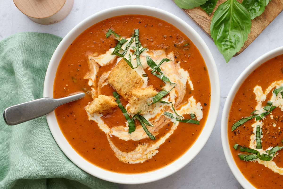 Two bowls of homemade tomato soup garnished with a few croutons and cream swirl, and basil leaves with a spoon dipped inside.