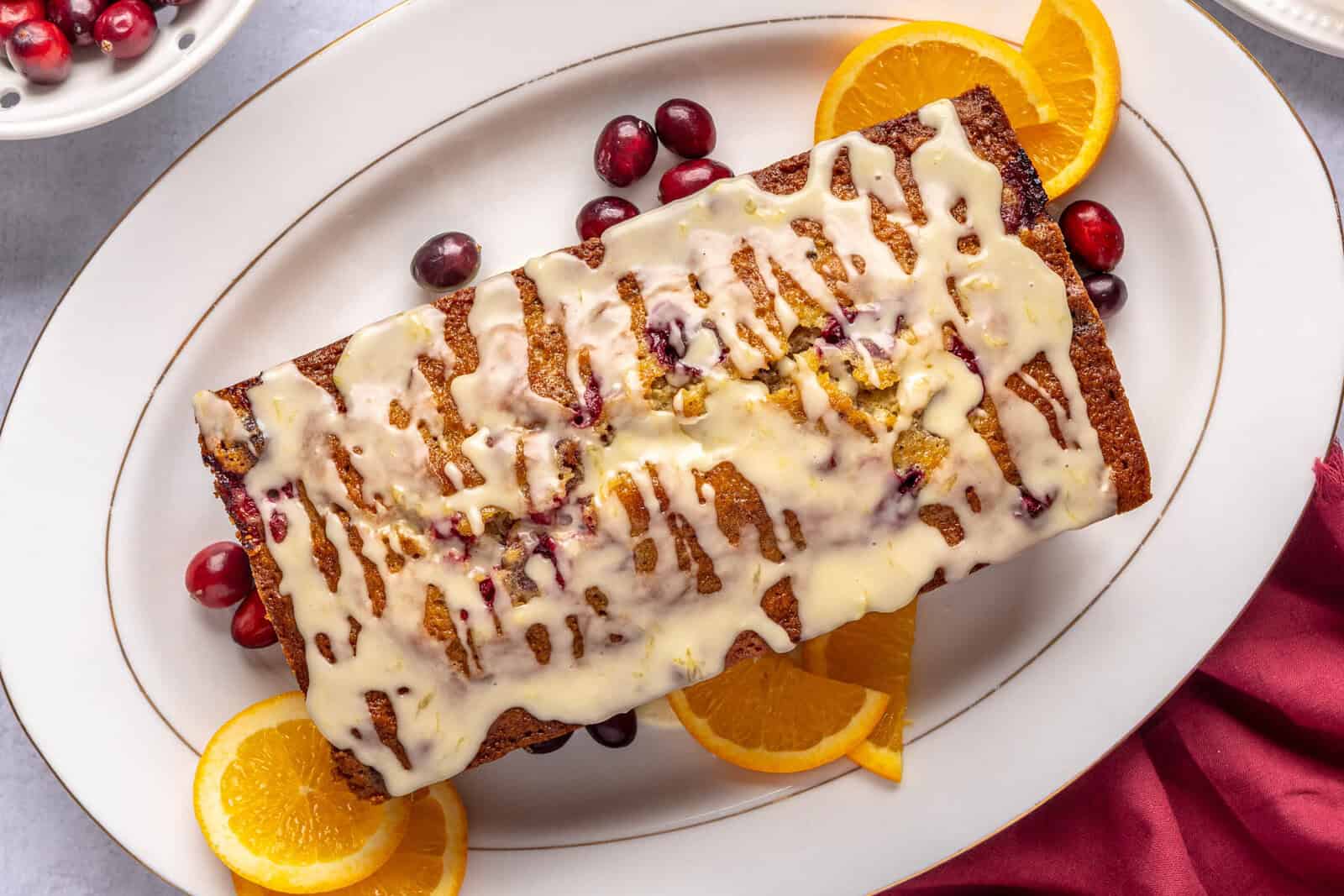 Cranberry orange bread with orange glaze on a narrow platter with a small dish of fresh cranberries and a few orange slices nearby.