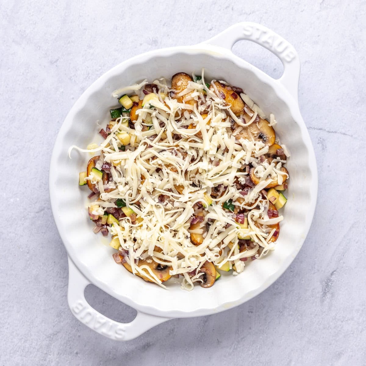 Cooked vegetables added to pie dish with cheese sprinkled on top for quiche recipe.