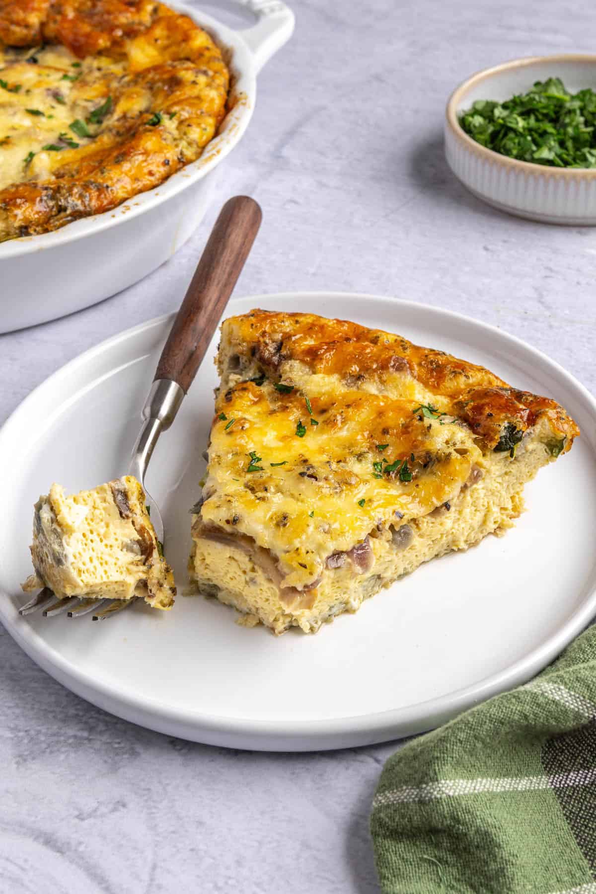 Slice of crustless breakfast quiche on a small plate with a bite taken and resting on fork with baking dish nearby.
