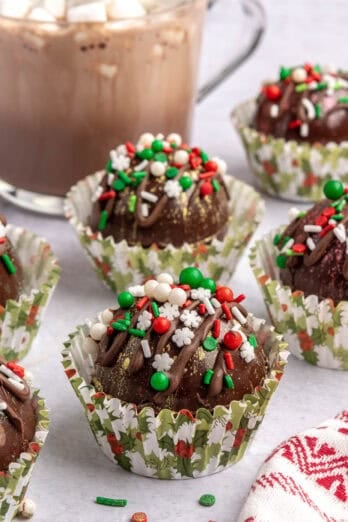 Hot cocoa bombs decorated with sprinkles in small paper liners with a cup of hot chocolate with marshmallows nearby.