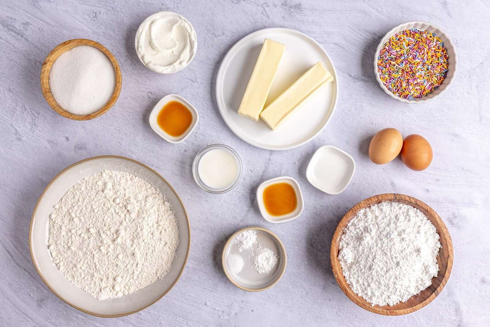 Ingredients for frosted lofthouse sugar cookies recipe: sugar, Greek yogurt, butter, vanilla, flour, eggs, salt, baking powder, baking soda, and sprinkles.