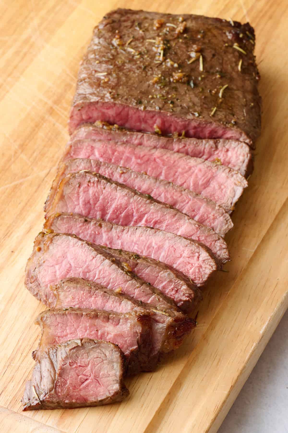 London broil on a carving board, sliced against the grain into thin ยผ inch slices about half way up, slices slightly spread out and laying forward to show medium rare temperature of the meat.