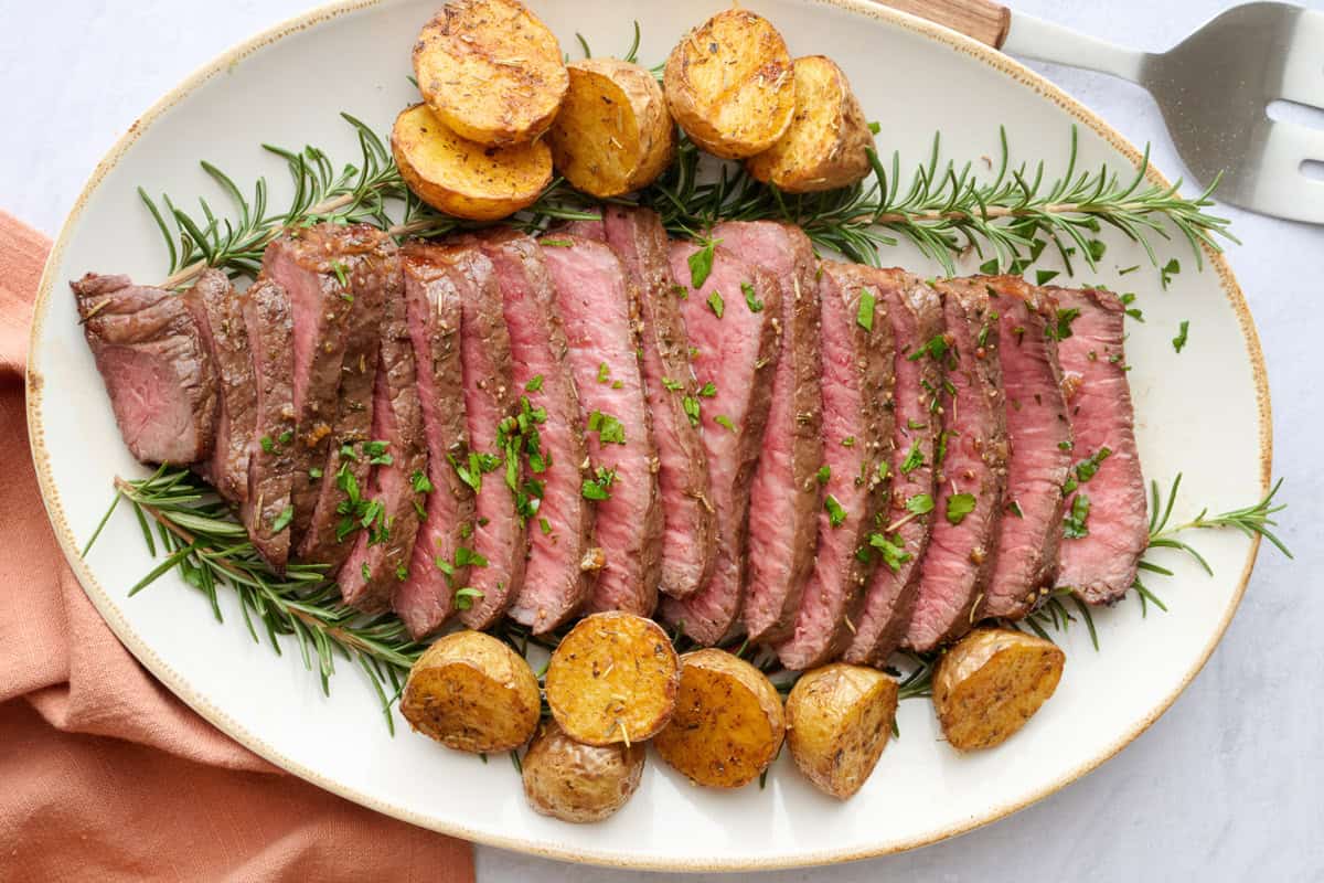 Thin slices of London broil on a platter with a few sprigs of fresh rosemary around and garnished with chopped parsley. Serving set resting nearby.