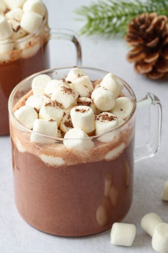 Two mugs of vegan hot chocolate topped with mini marshmallows and chocolate shavings,