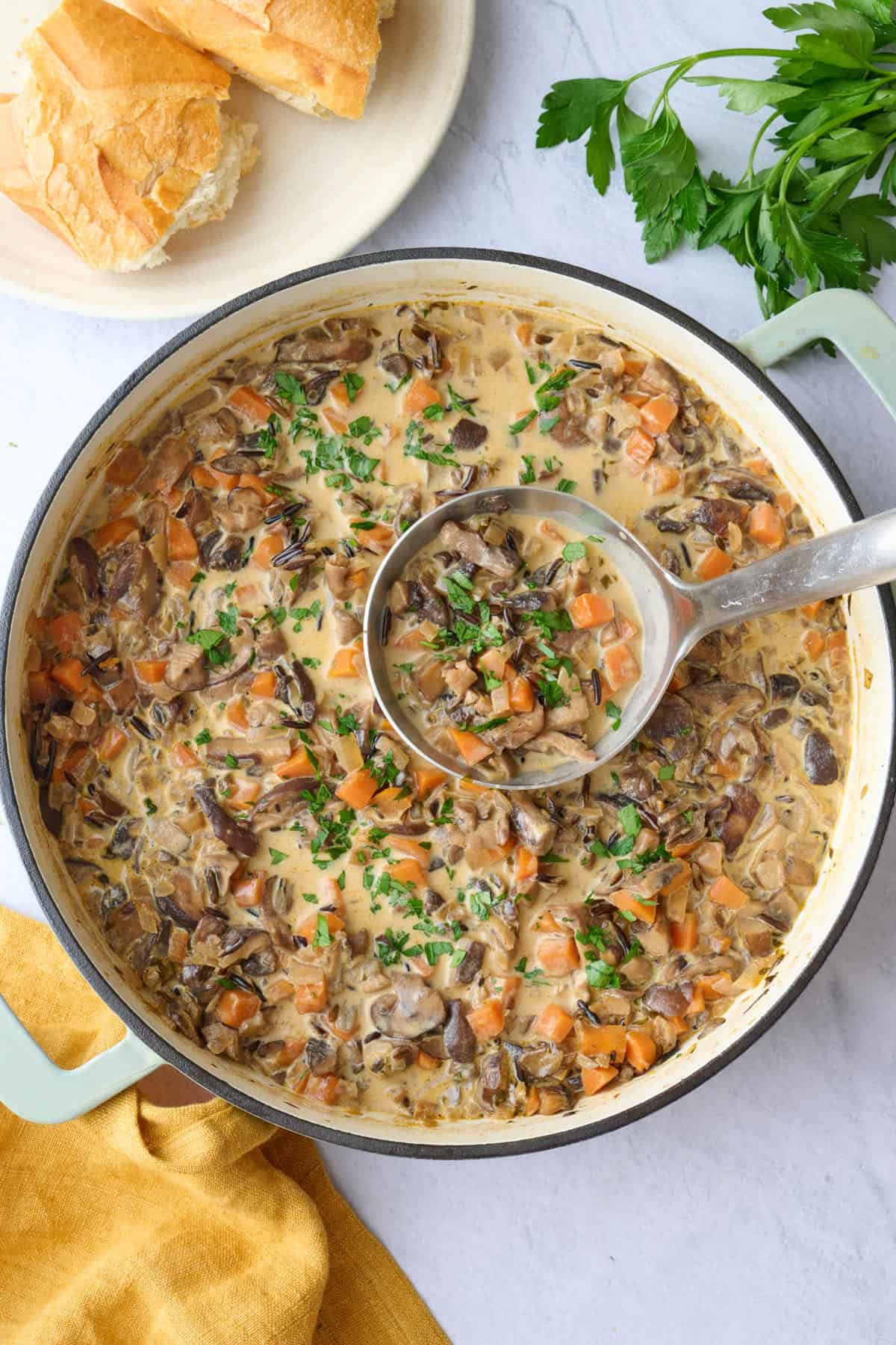 Wild rice mushroom soup in the pot garnished lightly with chopped parsley with a ladle lifting some up, plate of crusty bread nearby.