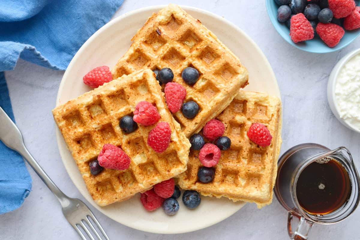 Oatmeal cottage cheese waffle on a plate topped with fresh berries with maple syrup nearby.