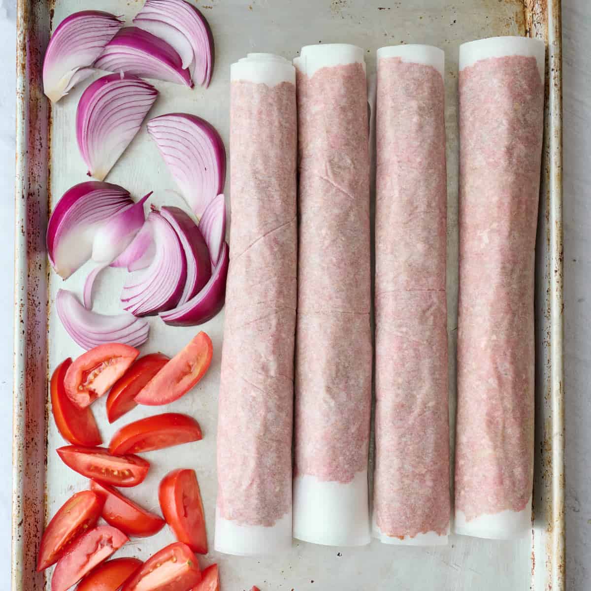 Four logs of homemade doner kebab meat mixture on a sheet pan with chunks of red onion and tomatoes before baking.