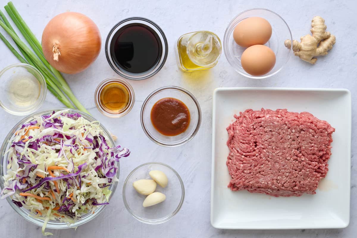Egg roll ingredients before prepping: rice vinegar, sesame seed oil, sriracha, rice vinegar, soy sauce, green onion, onion, coleslaw mix, garlic, oil, eggs, ginger, and ground beef.