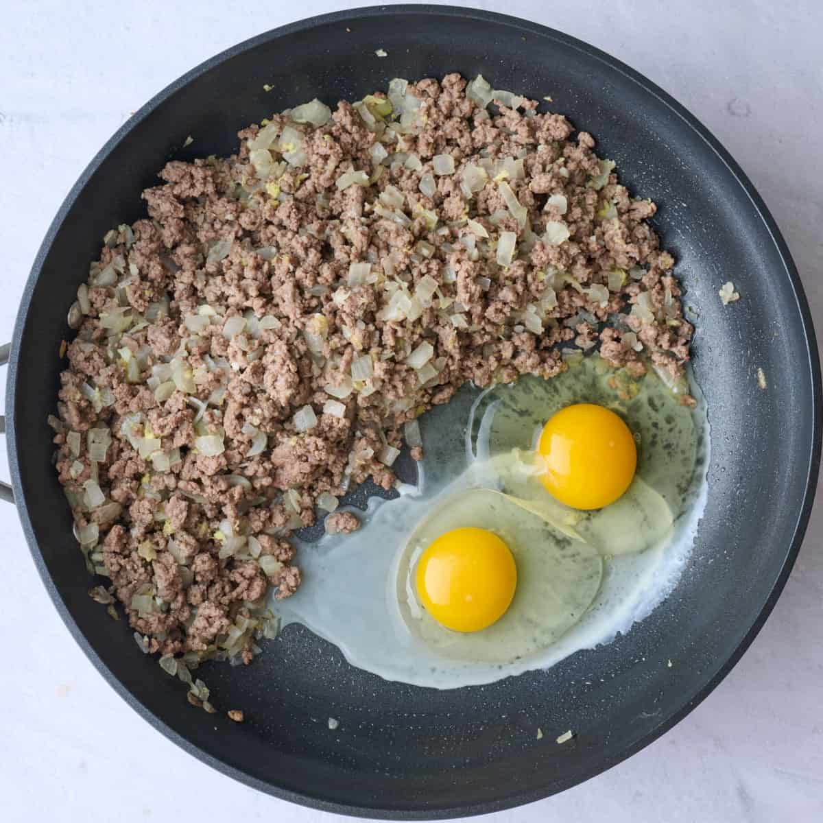Ground beef mixture after cooking with aromatics pushed to side with two eggs cracked onto the other side.