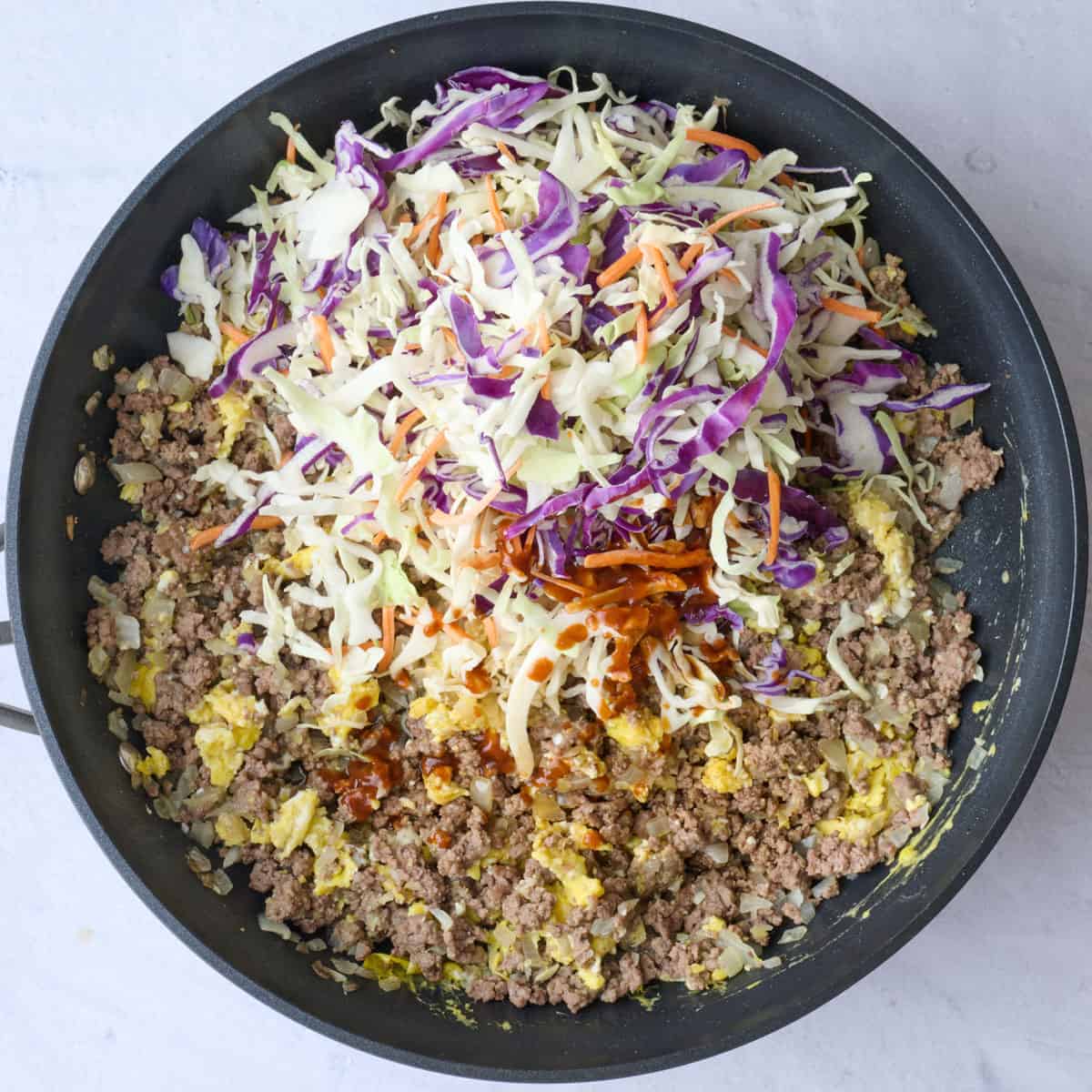 Egg rolling beef mixture in a skillet with coleslaw mix and sauces added on top before combining.