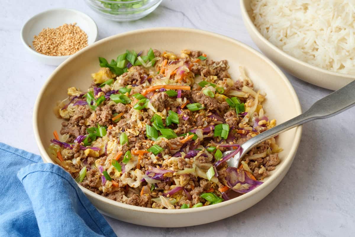 Beef egg roll in a bowl with coleslaw mix with dishes of white rice, sesame seeds and green onions nearby.