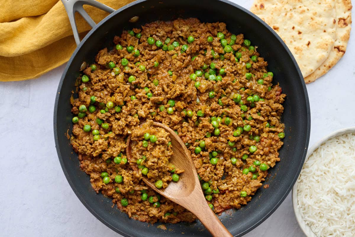 Classic keema curry recipe with seasoned ground beef and peas in a skillet with serving spoon dipped inside and a bowl of cooked rice and naan nearby.