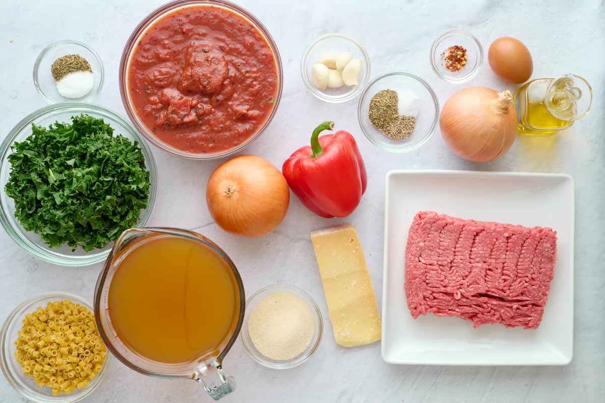Ingredients for soup recipe with homemade meatballs: kale, pasta, broth, crushed tomatoes, onion, red pepper, parmesan cheese, garlic, seasonings, egg, oil and ground beef.