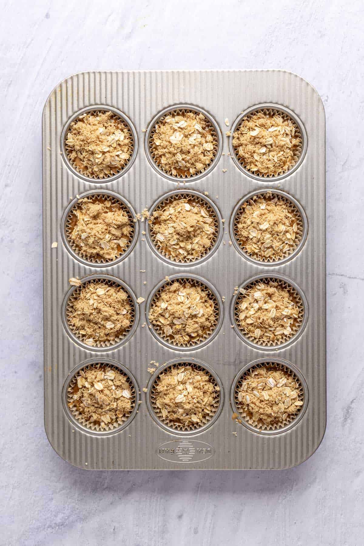 Oatmeal muffin batter divided into a 12 cup muffin pan with paper liners and oat topping sprinkled on top before baking .