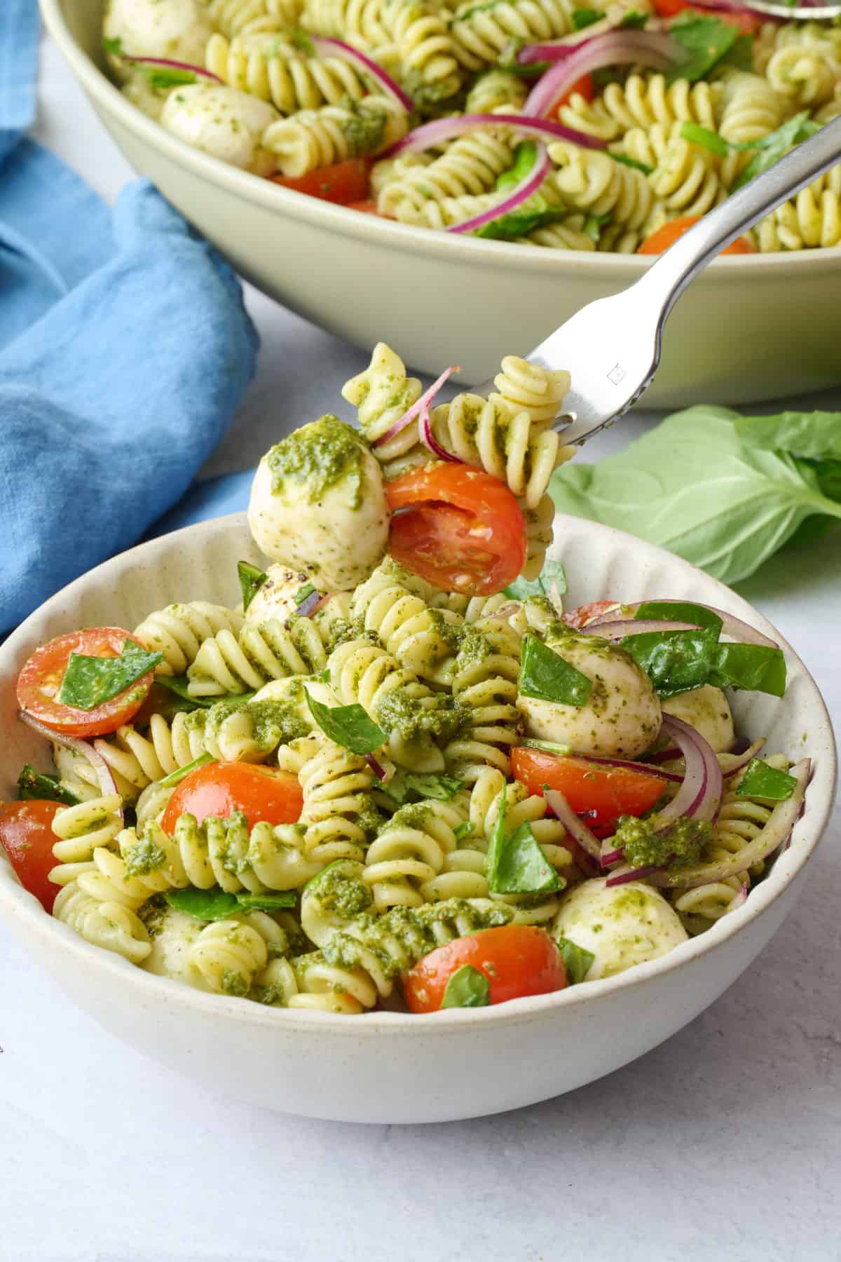 Best pesto salad with pasta, tomatoes, mozzarella, onions and spinach in a small bowl with a fork lifting up a bite.