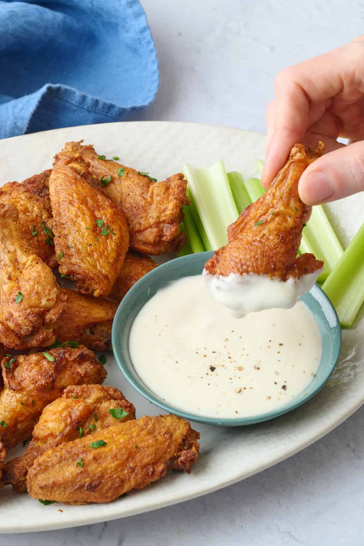 Chicken wing being dipped into a dish of ranch with more baked wings on platter.