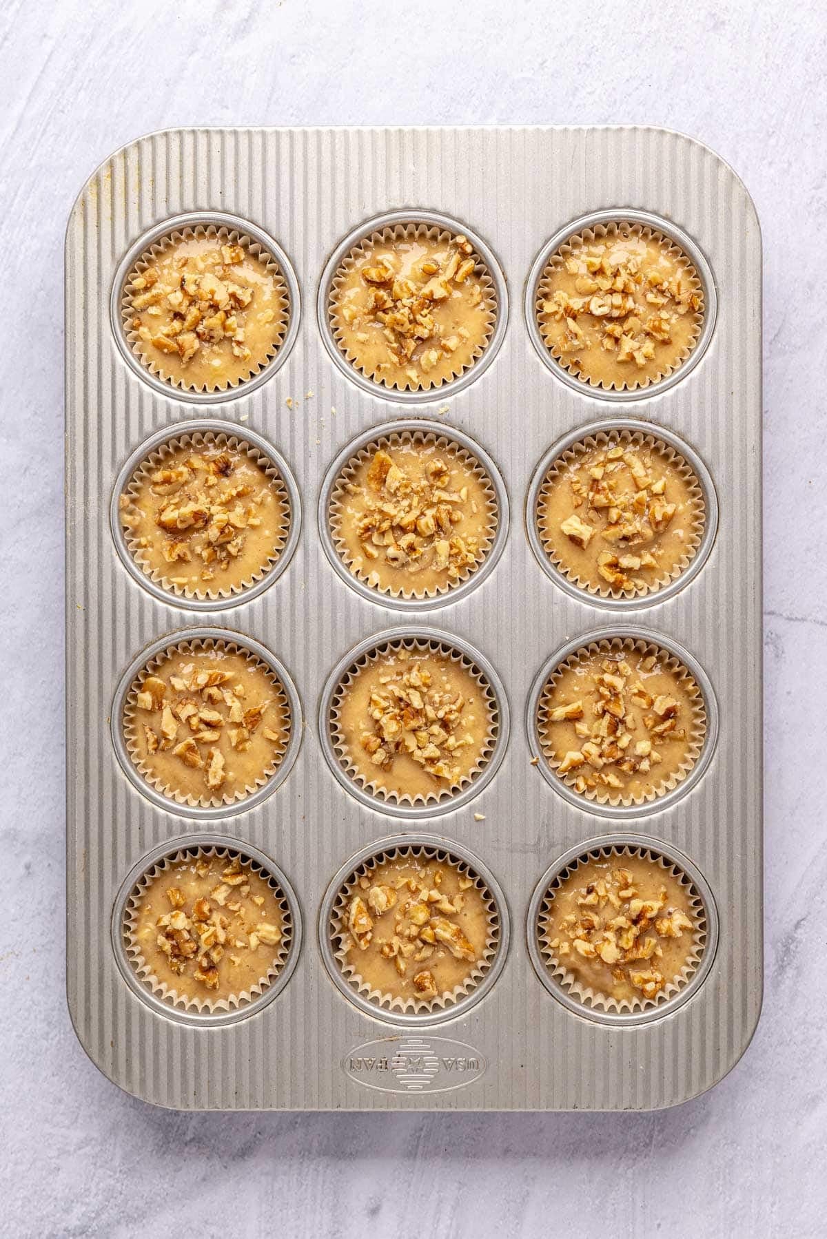 Banana muffin batter divided into a 12 cup muffin pan lined topped with walnuts before baking.
