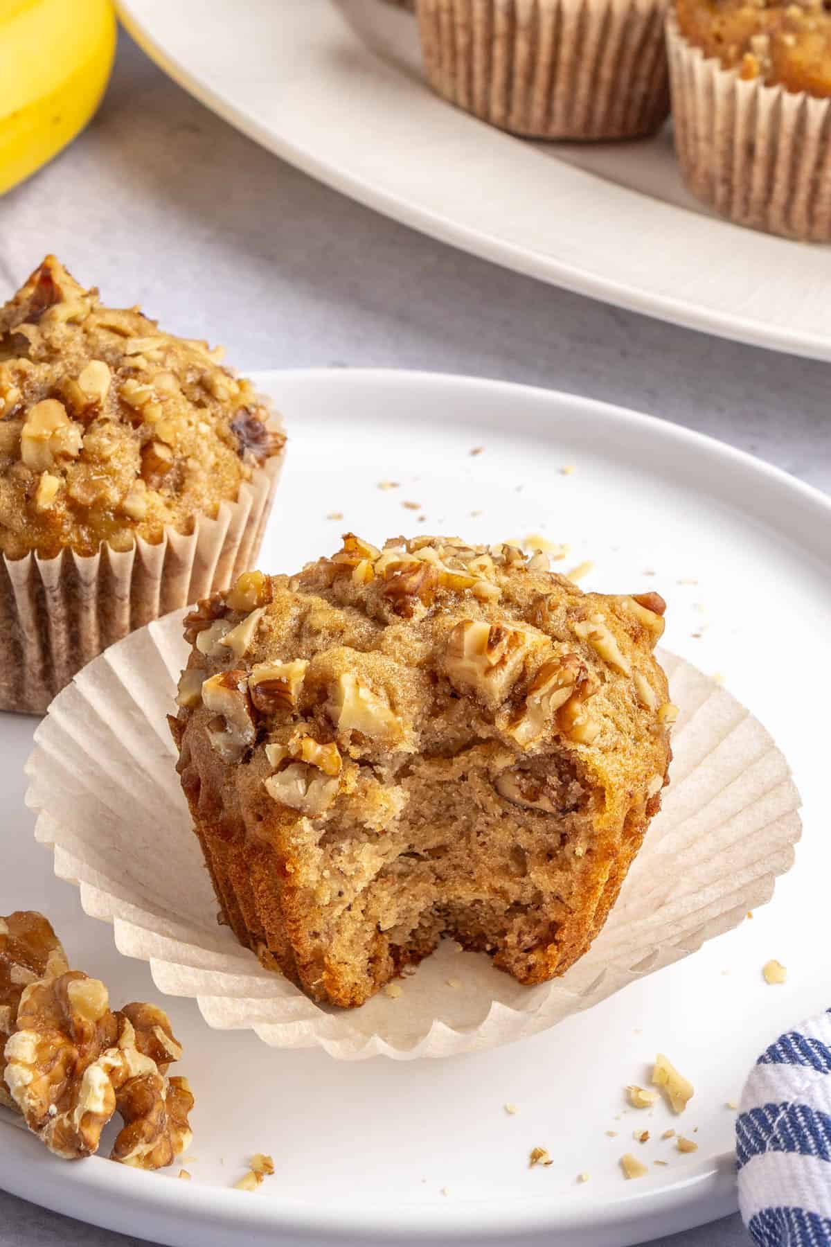 Banana walnut muffin on a paper liner after peeling back and a bite taken out of it to show soft inside texture.