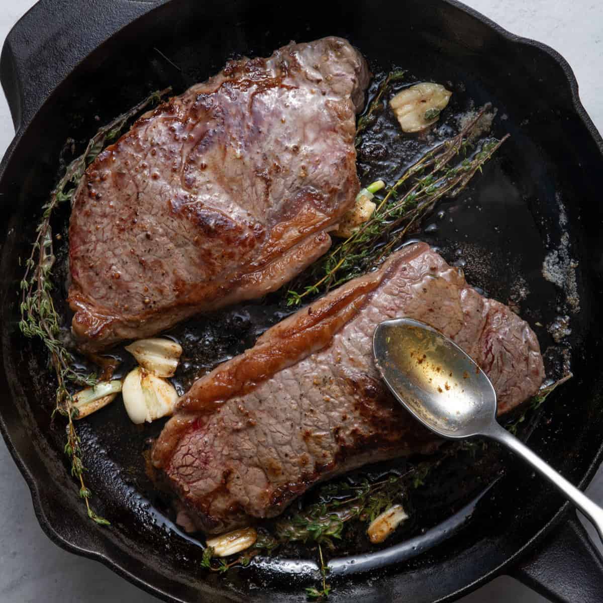 Spooning melted butter over steaks in cast iron skillet.
