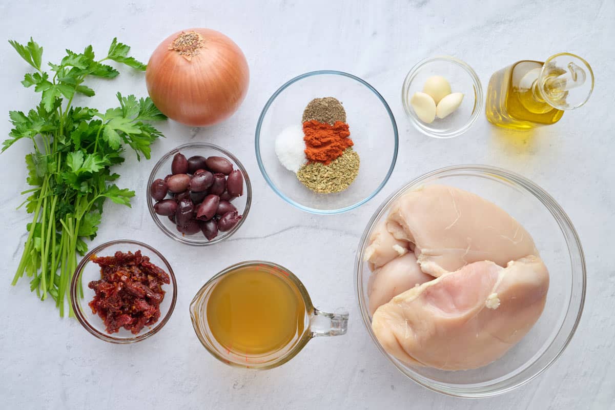 Ingredients for Mediterranean chicken recipe: parsley, onion, kalamative olives, sun dried tomatoes, chicken broth, seasoning blend, chicken breasts, garlic cloves and oil.