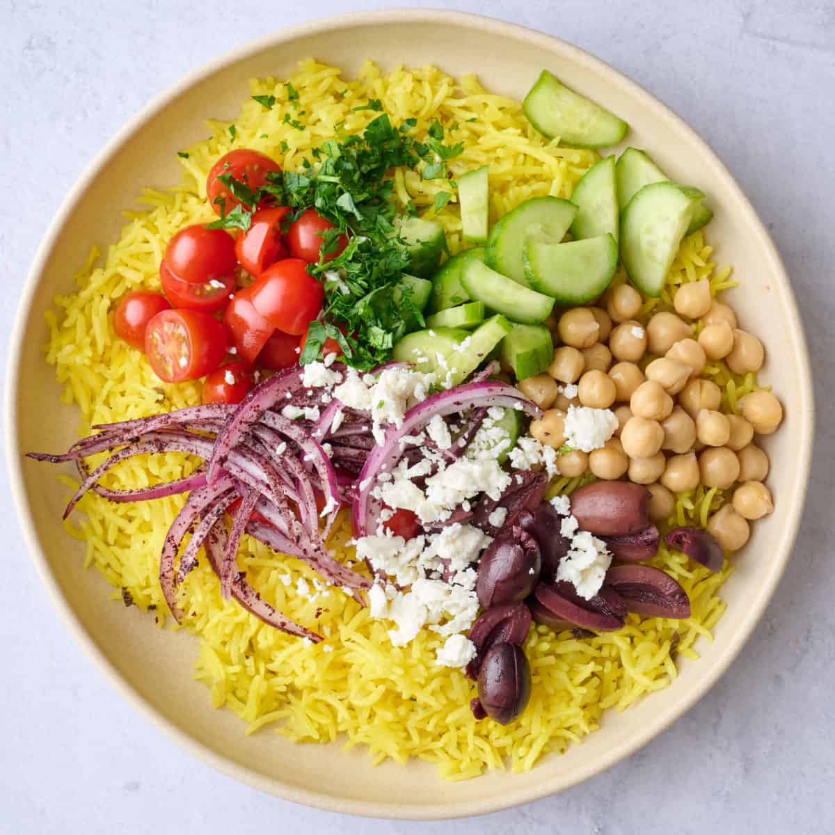 Halves cherry tomatoes, parsley, sliced Persian cucumbers, chickpeas, feta, sumac sliced red onions, and kalamata olives added on top of rice in a bowl.