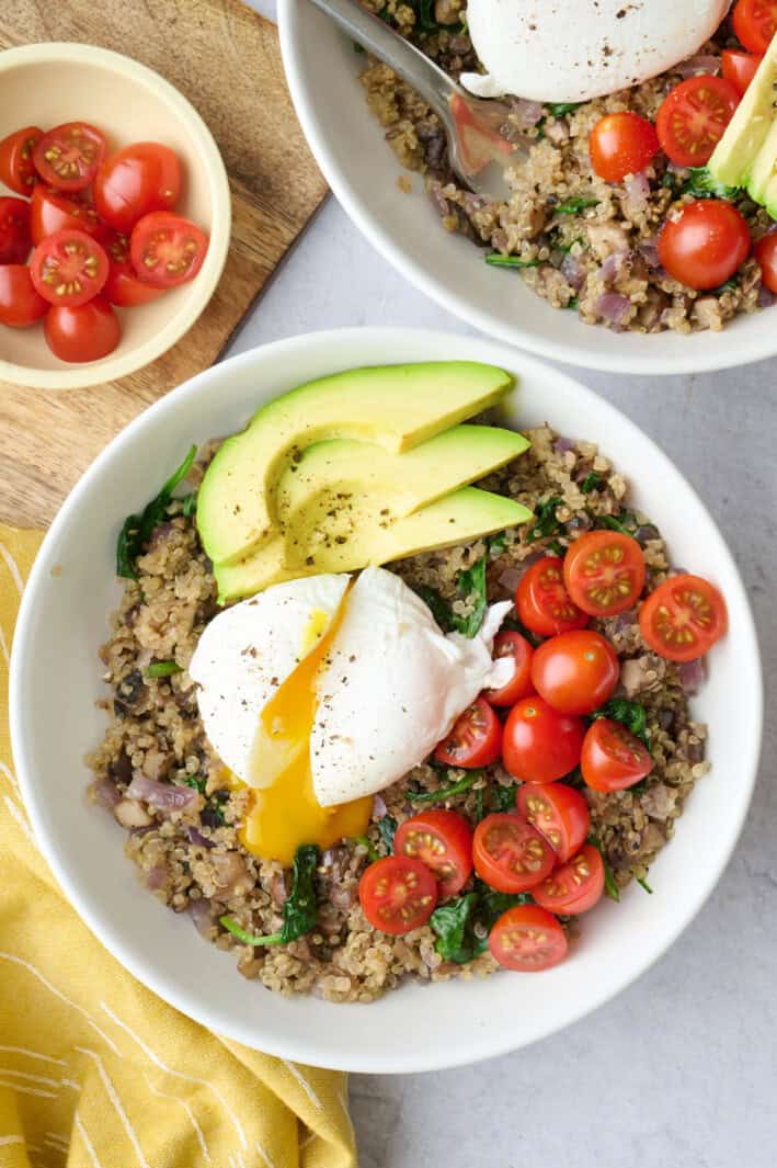 Two quinoa breakfast bowls topped with a poached egg, broken and running out, plus sliced avocado, and sliced cherry tomatoes.