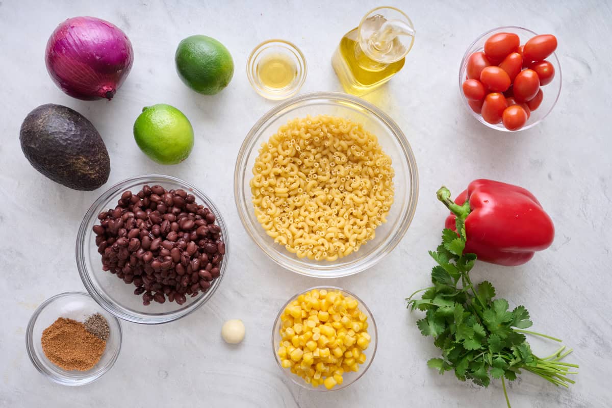 Ingredients for cold pasta salad: black beans, avocado, red onion, lime, seasonings, garlic, corn, pasta, oil, tomatoes, red pepper, and cilantro.