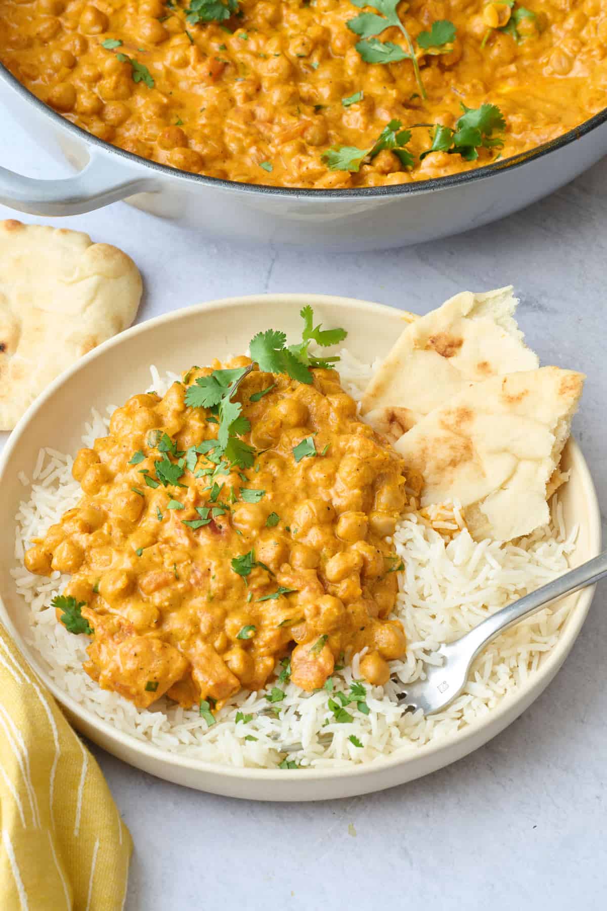 Serving of chickpea curry next to rice in a shallow bowl garnished with cilantro leaves, torn naan tucked into bowl with skillet of curry nearby.