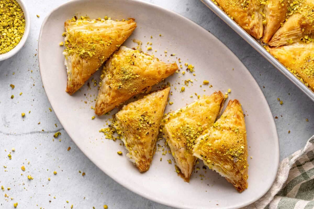 A few Shaabiyat on a long narrow platter garnished with crushed pistachio, baking dish with more still in square shape and a small dish of crushed pistachios nearby.